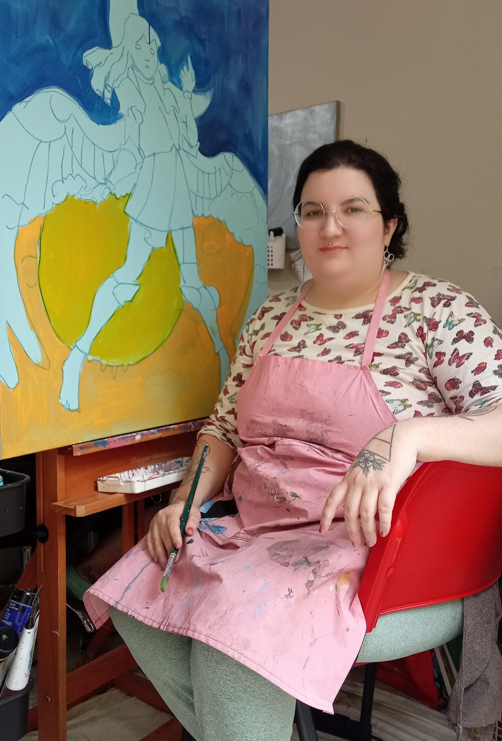 Foto da Artista no ateliê. Sentada, vestindo uma blusa de borboletas e um avental rosa, segurando um pincel na mão. Ela tem óculos, piercing medusa e brincos prateados, e um sorriso confiante no rosto. Atrás dela está seu quadro incompleto de um anjo.